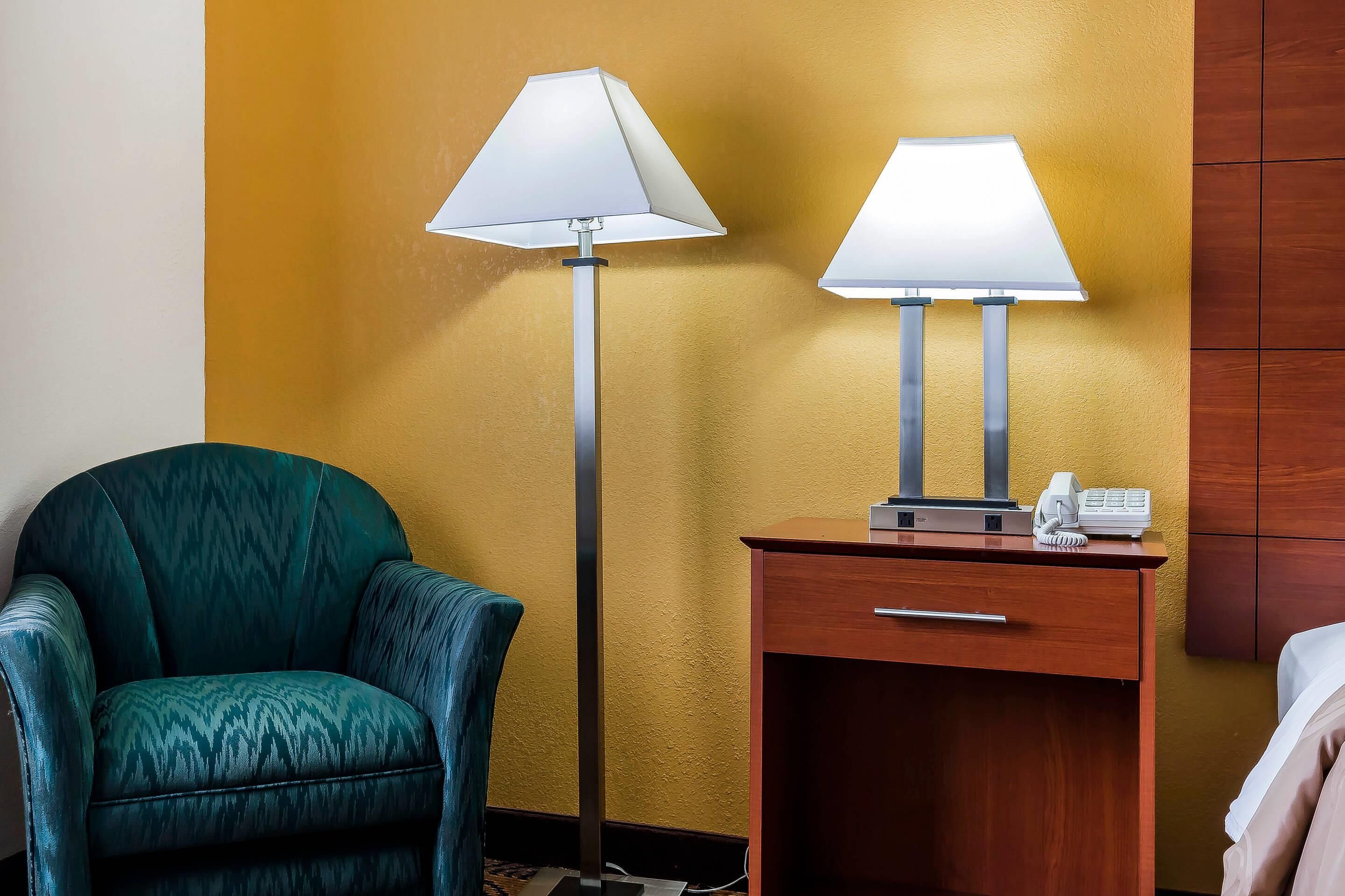 A hotel room with a teal armchair, a wooden nightstand with a phone, and two floor lamps.