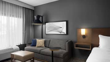 A modern hotel room with a grey sofa, a small round table, a bedside table with a lamp, a framed abstract artwork, and a large window with sheer curtains.