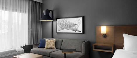 Ein modernes Hotelzimmer mit grauem Sofa, einem kleinen runden Tisch, einem Nachttisch mit Lampe, einem gerahmten abstrakten Kunstwerk und einem großen Fenster mit durchsichtigen Vorhängen.
