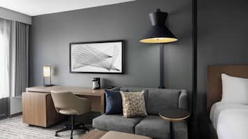 A modern hotel room with a grey sofa, a small round table, a desk with a chair, a bed, a framed abstract artwork, and a floor lamp.