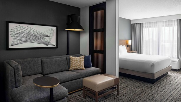 A modern hotel room with a grey sofa, a round table, a bed with white linens, a large abstract painting, a floor lamp, and a window with curtains. - Hyatt Place Tempe Phoenix Airport (Tempe)