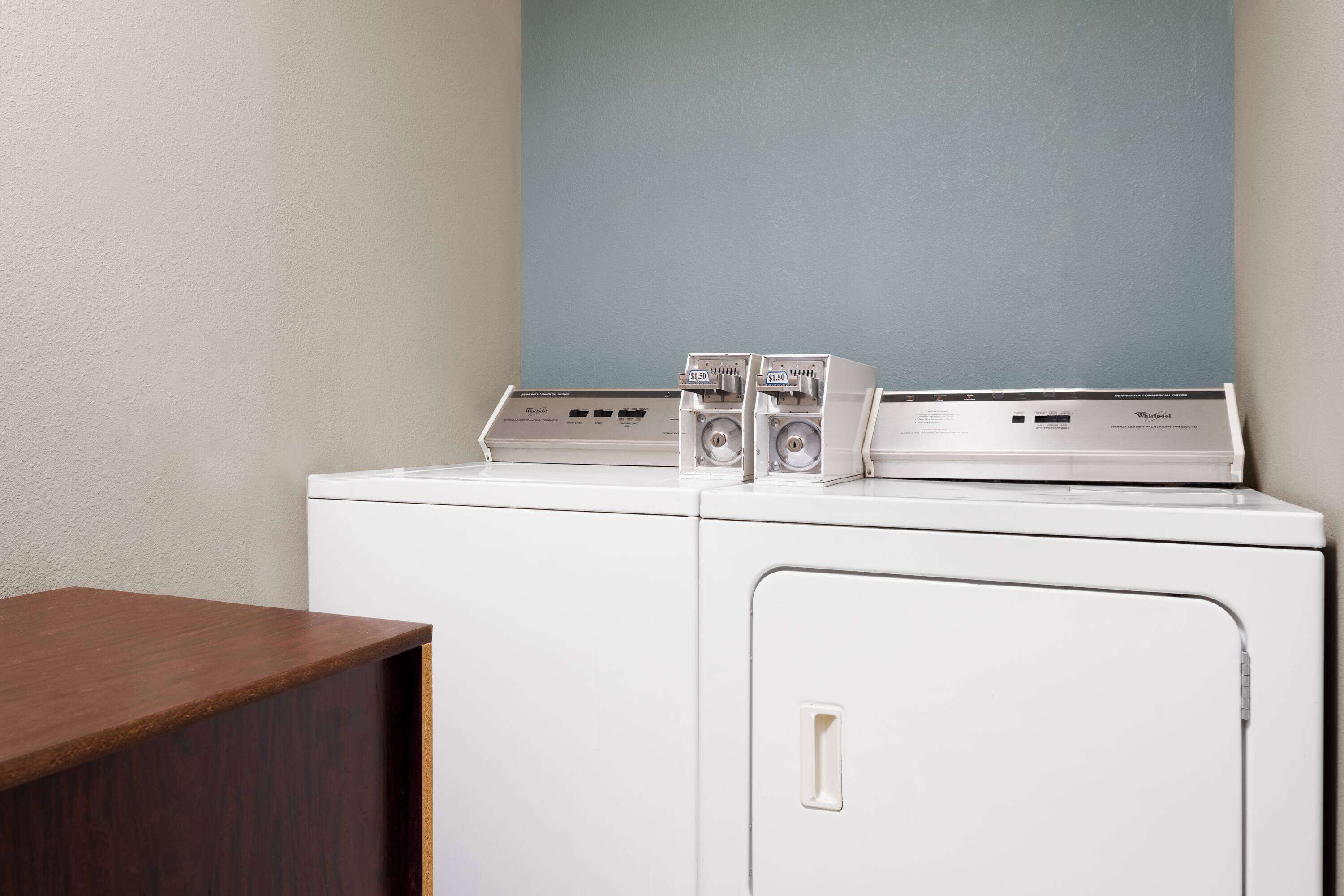 A white laundry machine with a control panel and two coin slots, positioned against a light blue wall.
