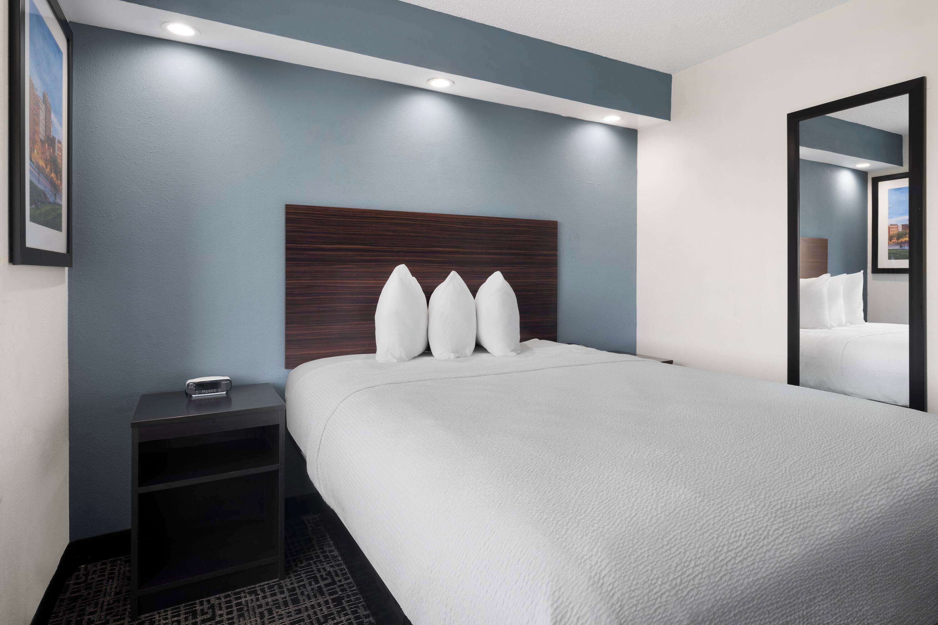 Quarto de hotel com uma cama grande, uma mesa de cabeceira, um espelho e cabeceira fixada na parede.