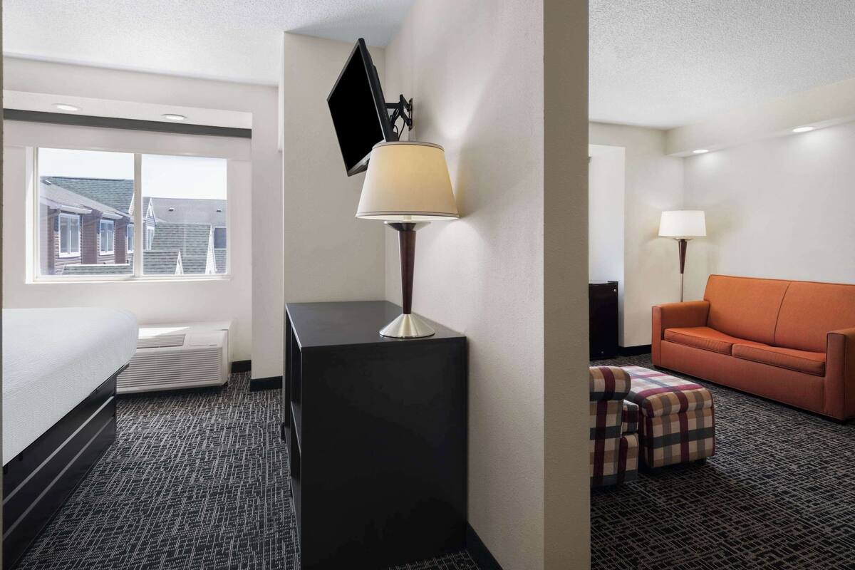 A hotel room with a bed, a desk, a lamp, a TV, an orange sofa, and a checkered ottoman.