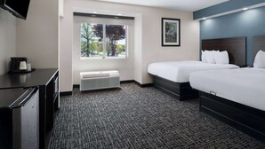 A hotel room with a large bed, a smaller bed, a desk with a flat-screen TV, a heater, and a window with a view of trees. - Baymont by Wyndham Boardman (Boardman)