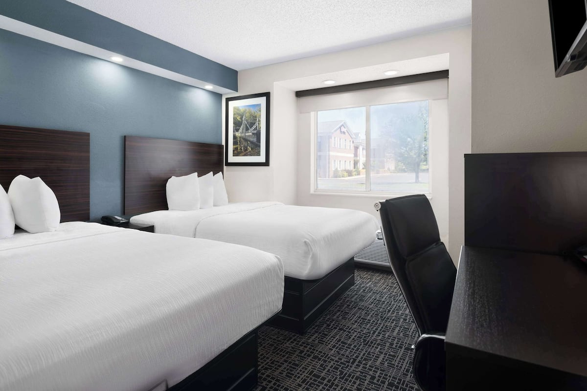A hotel room with two beds, a desk, a chair, a TV, and a window with a view of a building.