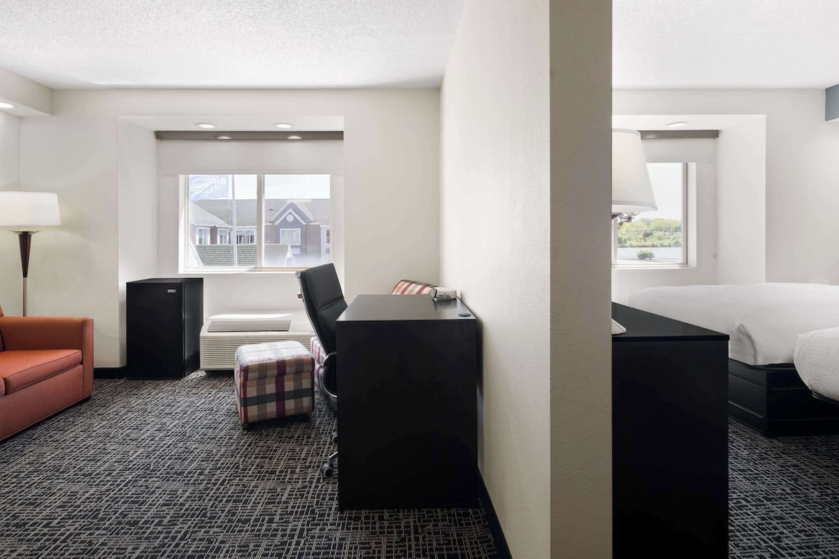 A hotel room with a bed, a desk, a chair, a red sofa, a small table, and a window with a view of buildings.