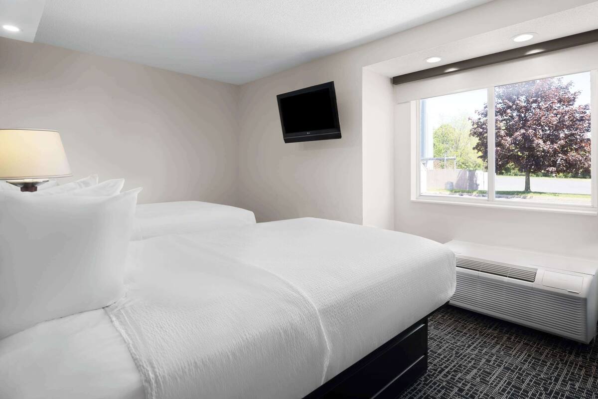 A hotel room with two beds, a flat-screen TV mounted on the wall, a window with a view of trees, and a wall-mounted air conditioning unit.