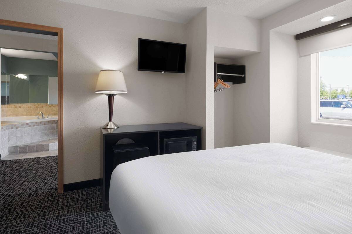 A hotel room with a bed, a TV mounted on the wall, a desk with a lamp, and a window.