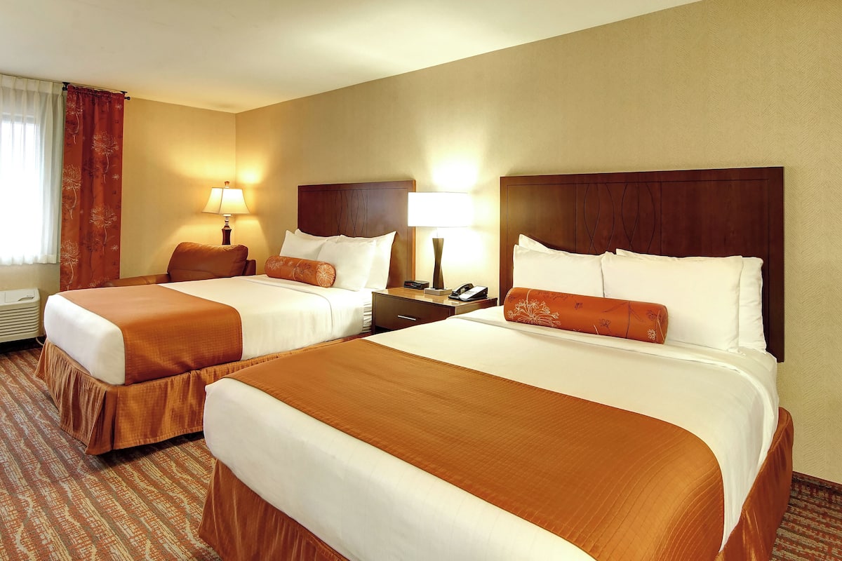 A hotel room with two beds, each with a brown bedspread, white pillows, and a wooden headboard. There is a nightstand with a lamp and a phone between the beds.