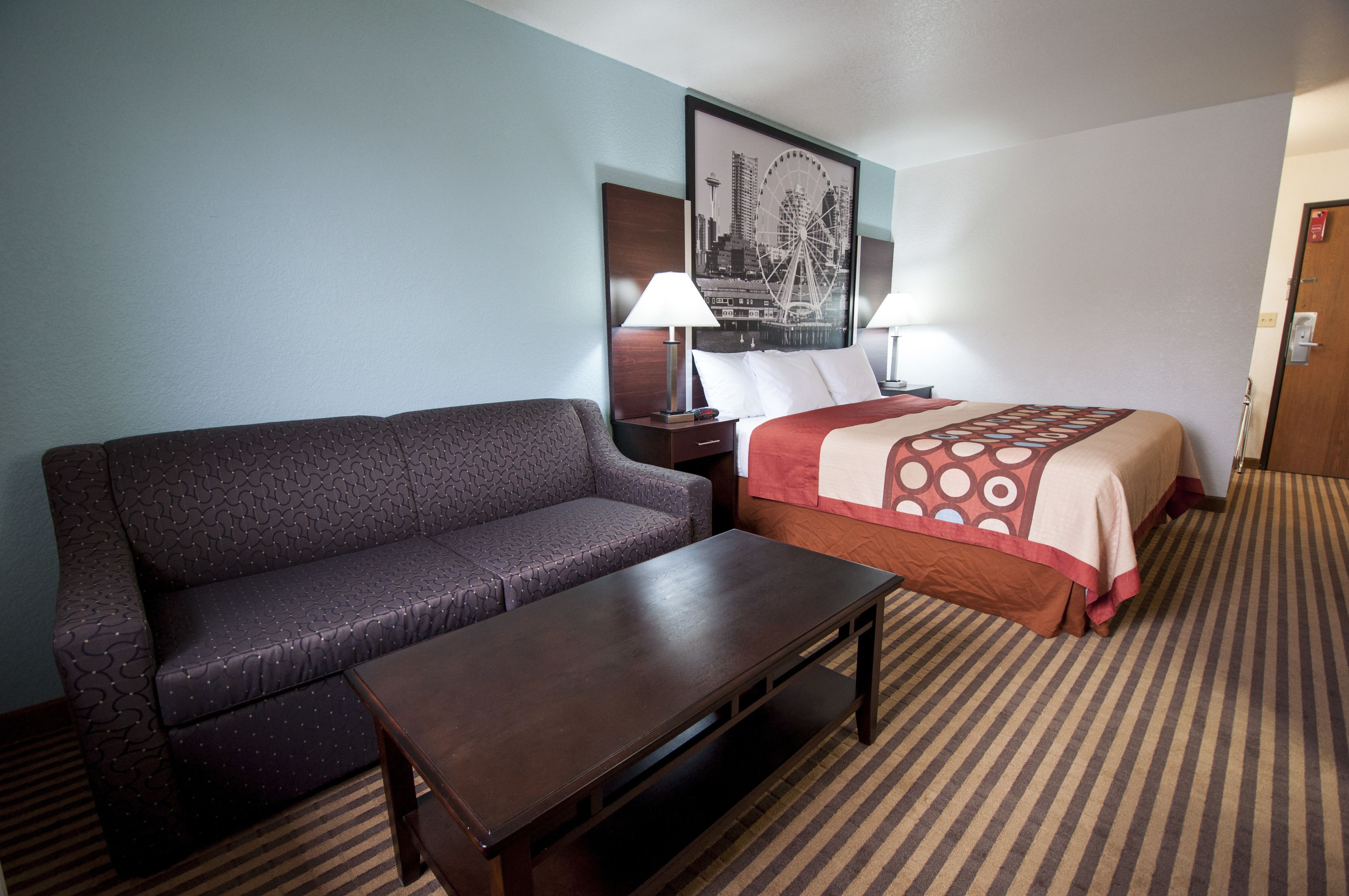 A hotel room with a sofa, a bed with a patterned bedspread, a nightstand with a lamp, and a framed cityscape picture on the wall.