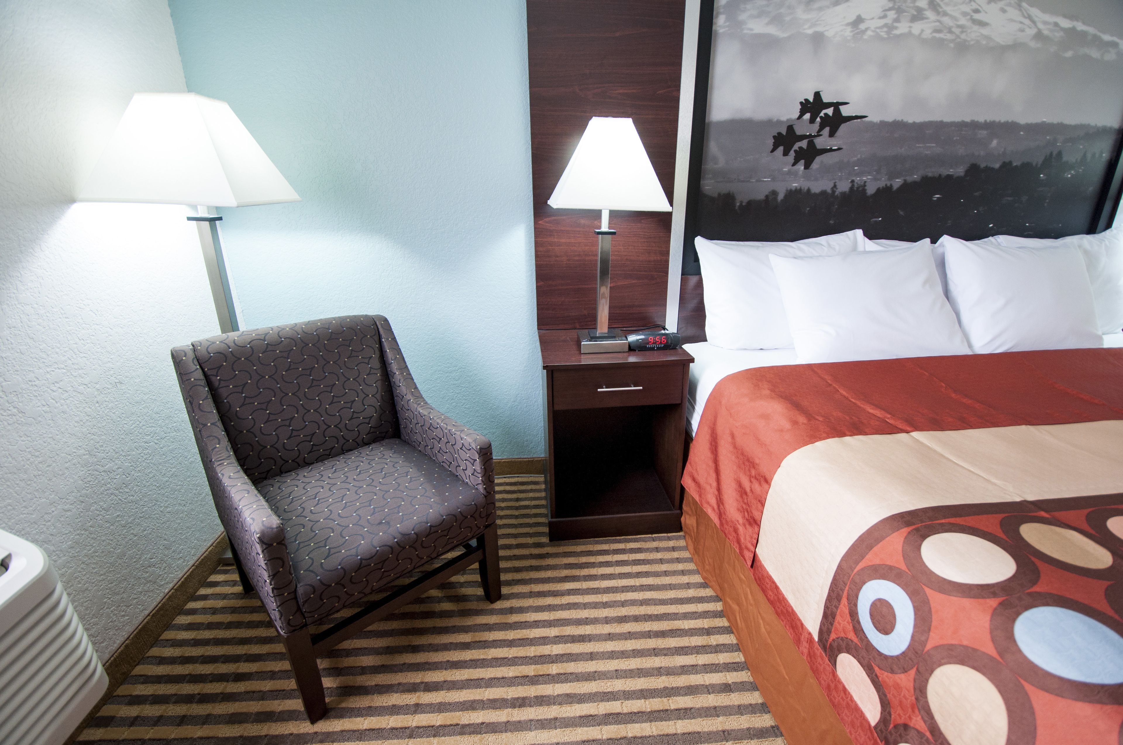A hotel room with a bed, a chair, a nightstand with a lamp, and a wall with a mountain view mural.