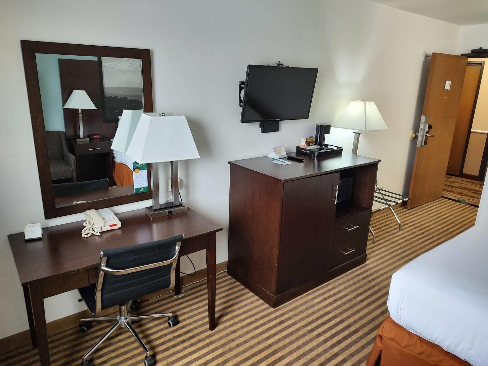 Habitación de hotel con una cama, un escritorio con silla, teléfono, televisor y espejo.