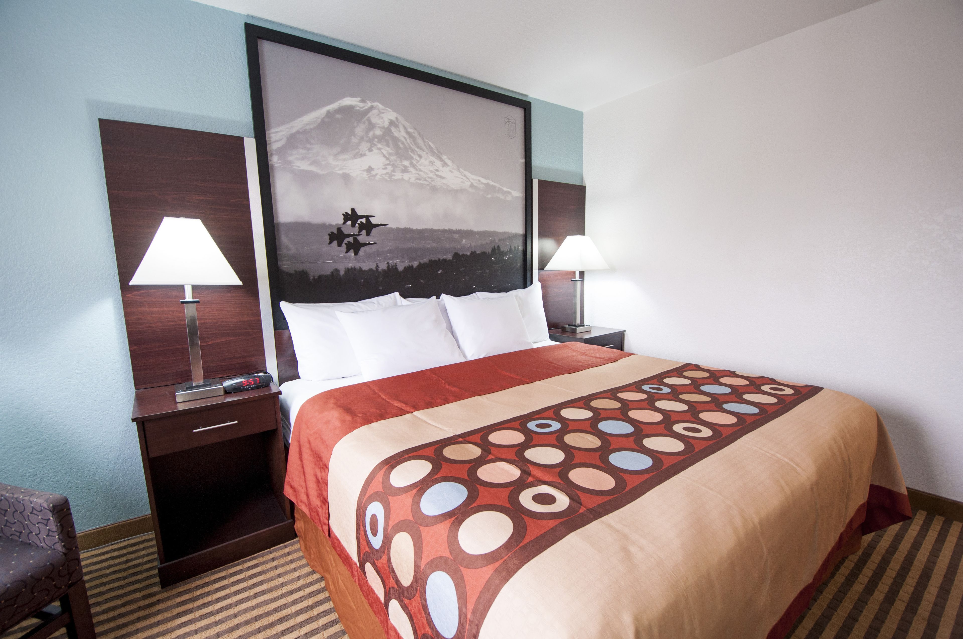A hotel room with a bed, two bedside tables, a nightstand, a chair, and a large framed picture of a mountain.