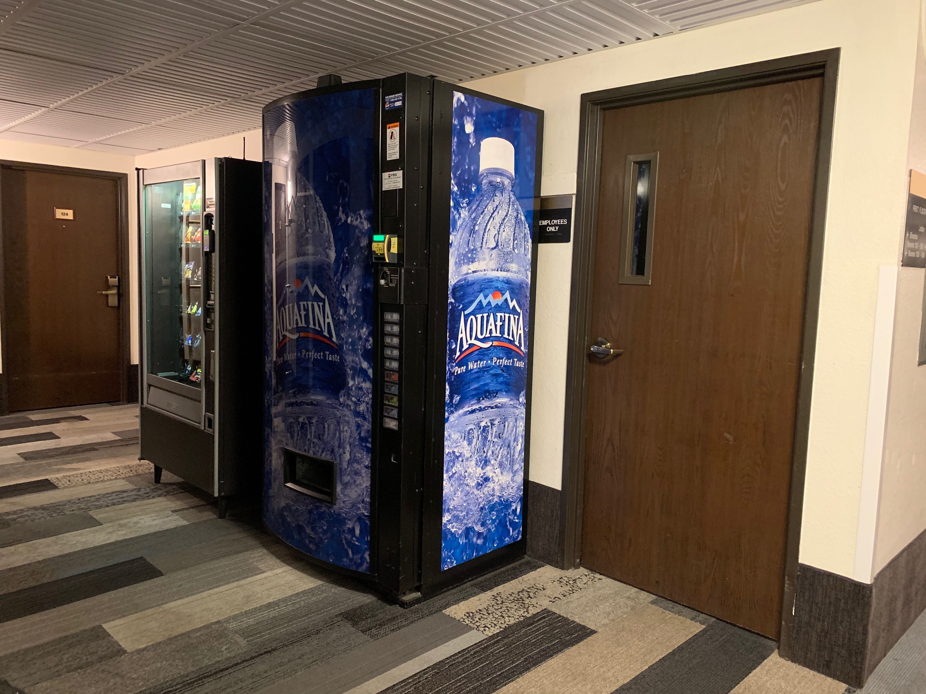 Un distributeur automatique de bouteilles d’eau Aquafina, une porte avec un panneau indiquant un numéro, et un sol recouvert de moquette.