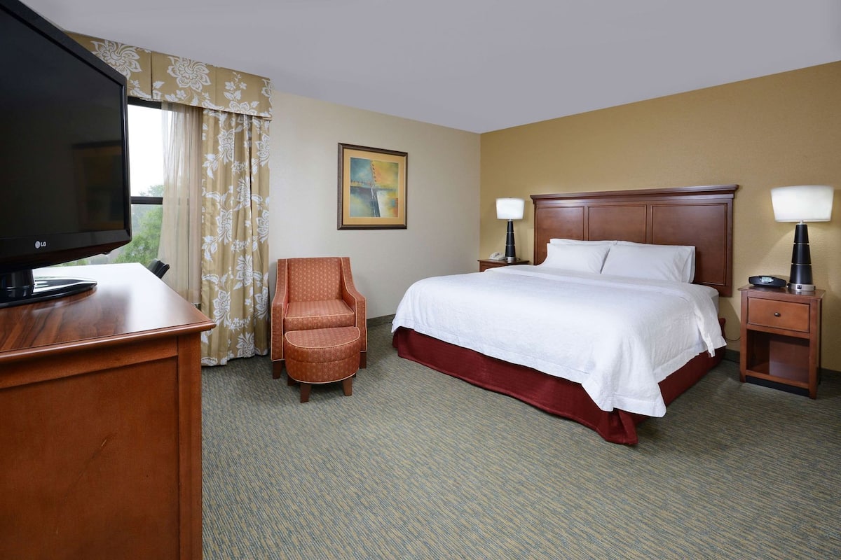 A hotel room with a bed, a chair, a desk with a TV, and a nightstand with a lamp.