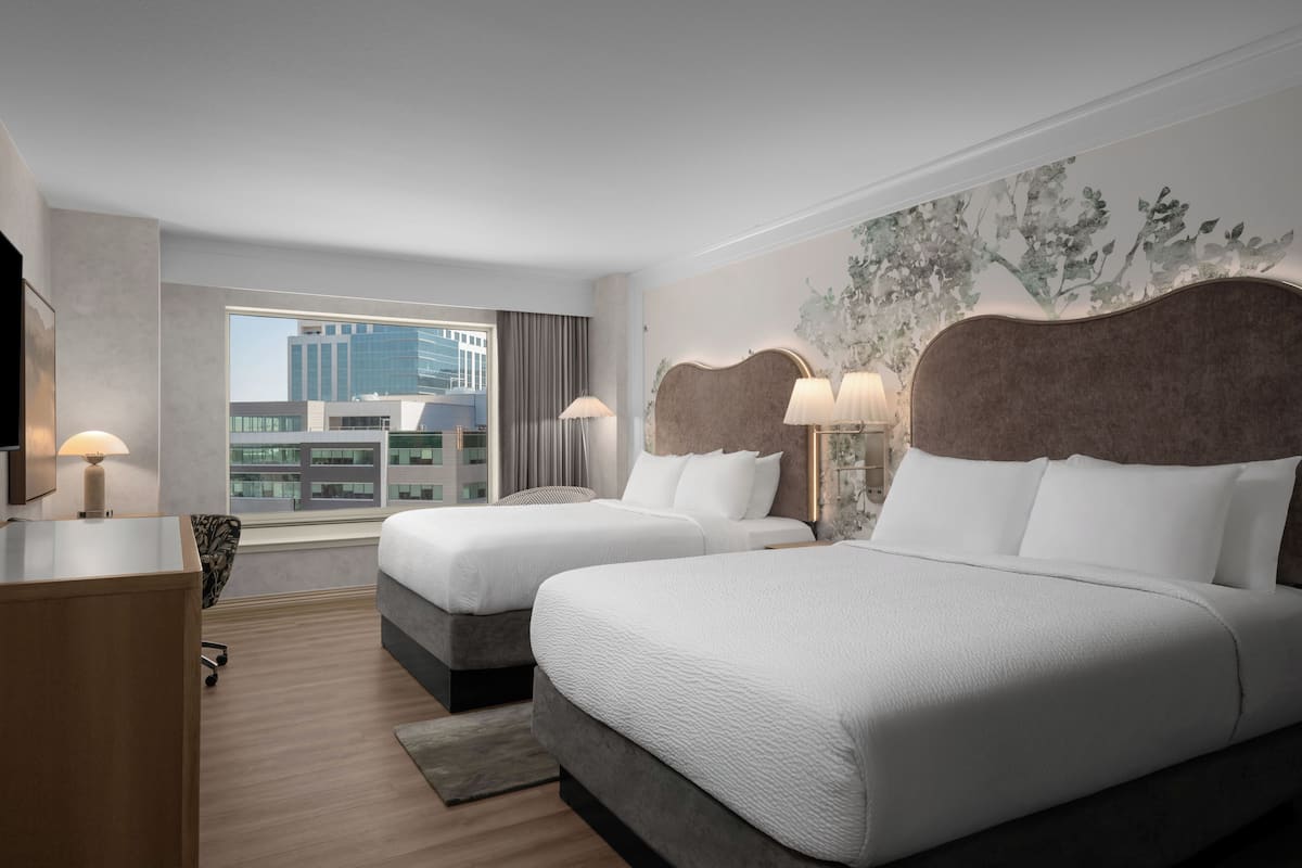 A hotel room with two beds, a desk, and a large window offering a city view.