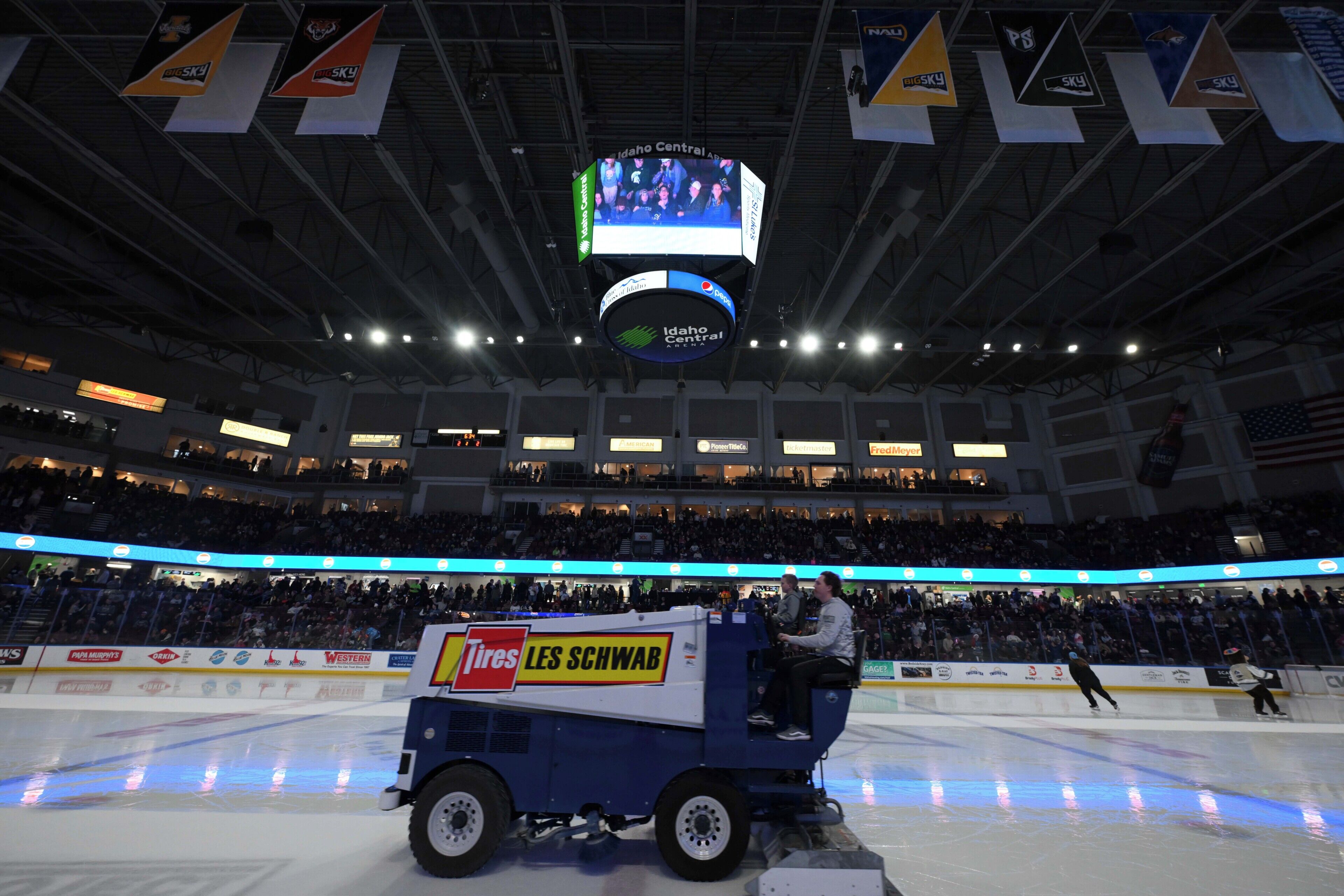 An indoor ice rink with a scoreboard, advertisements, and a person on a cleaning vehicle.