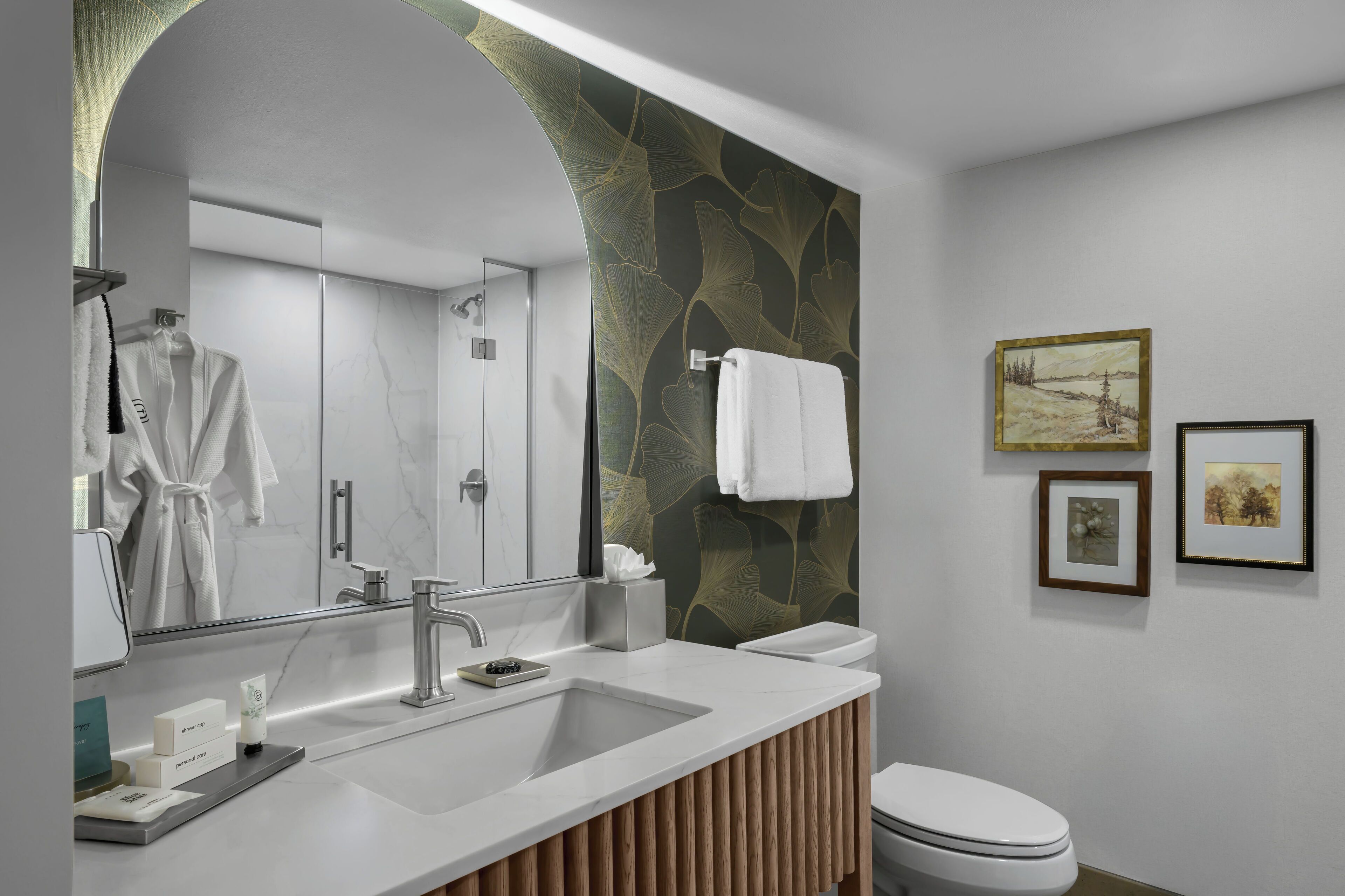 A modern bathroom with a large mirror, a sink, and a toilet. There are towels hanging on a rack, and a shower is visible in the background.
