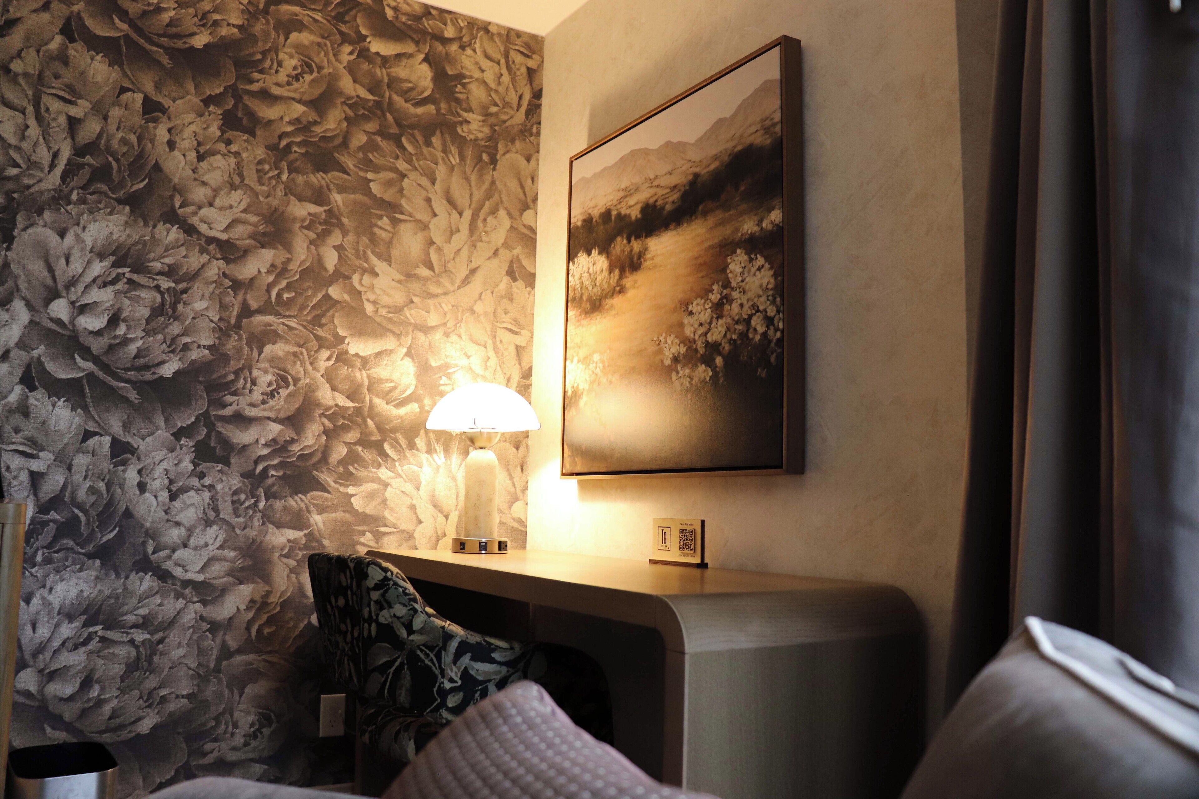A hotel room with a floral wallpaper, a mounted painting, a bedside table with a lamp, and a bed with pillows.