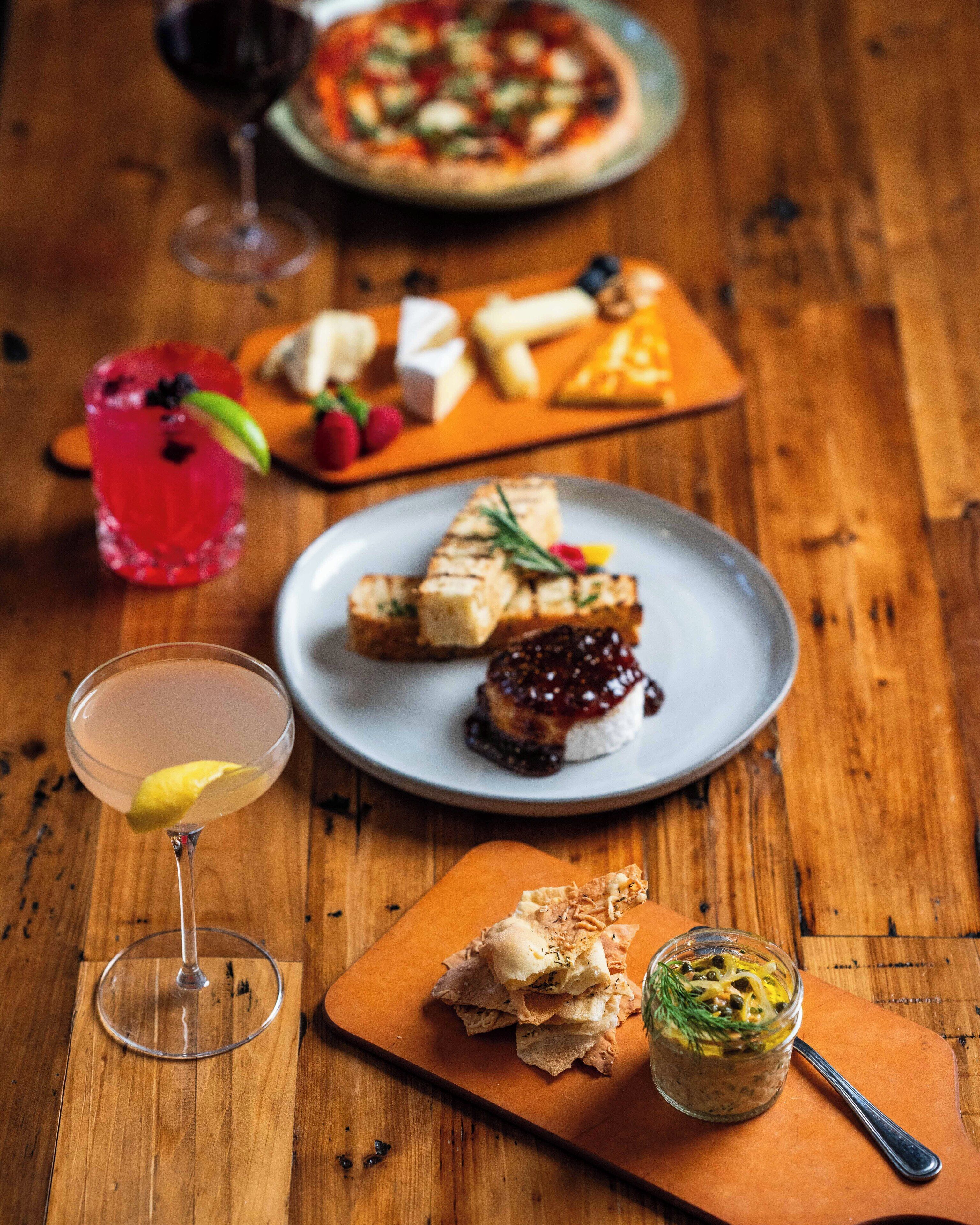 Une table en bois sur laquelle on trouve divers aliments, notamment une pizza, du fromage, des fruits, un verre à martini et un pot de pâte à tartiner.