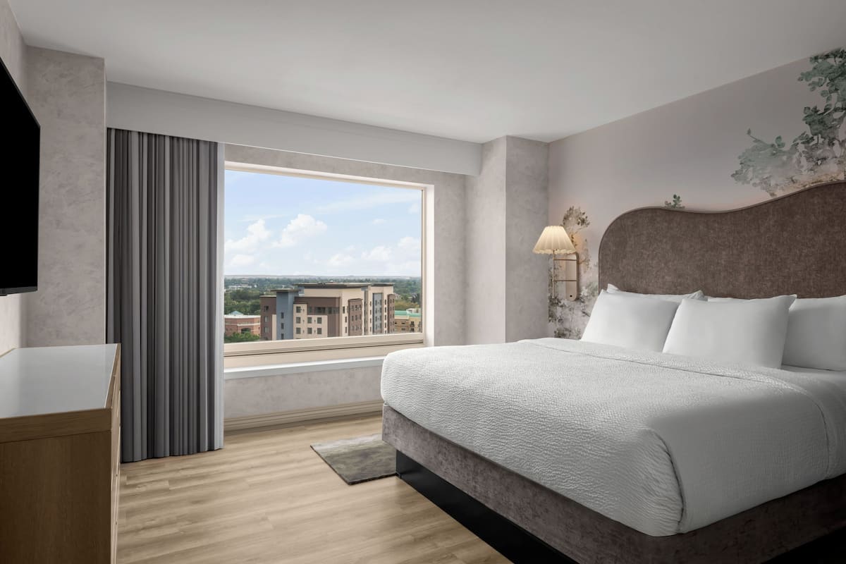 A modern hotel room with a large bed, a desk, a window with a view of buildings, and a lamp on the wall.