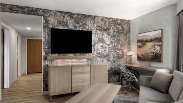 Un soggiorno moderno con TV a schermo piatto montata su una parete a motivi floreali, un tavolino di legno, un divano grigio e una scrivania con lampada.