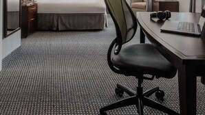 A hotel room with a bed, a desk with a laptop and headphones, a chair, a bedside table with a lamp, and a large window with curtains. - Club Quarters Hotel Faneuil Hall, Boston (Boston)