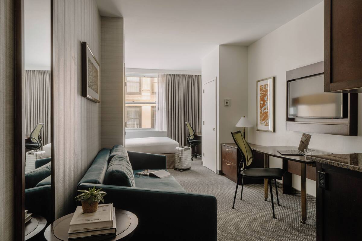 Een hotelkamer met een bed, een bureau, een stoel, een bank, een bijzettafeltje met een plant en een televisie.