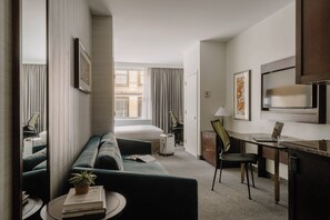 A hotel room with a bed, a desk, a chair, a sofa, a small table with a plant, and a TV.