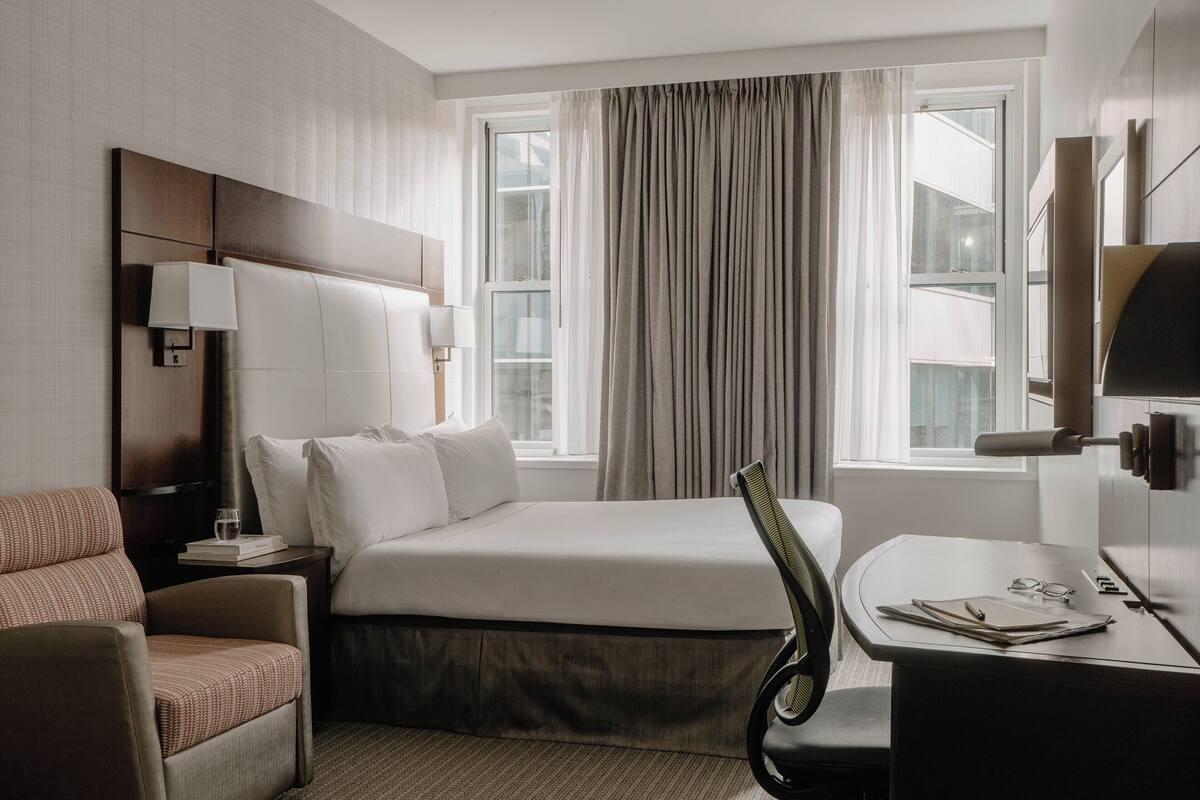 Een hotelkamer met een bed, een bureau, een stoel, een lamp en een raam met gordijnen.