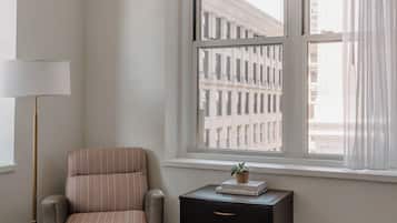 Une chambre d’hôtel avec un lit, une chaise, une table de chevet, une lampe et une fenêtre donnant sur des immeubles.