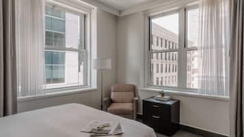 Una habitación de hotel con una cama grande, una silla, una mesita de noche y dos ventanas con cortinas.