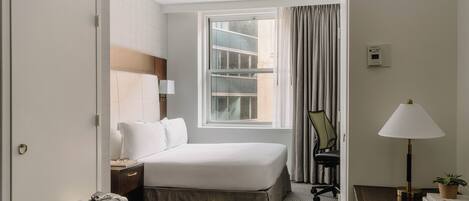 A hotel room with a bed, a desk with a lamp, a chair, a window with curtains, and a wall-mounted thermostat.