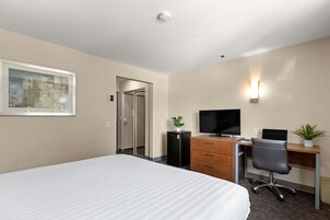 Habitación de hotel con una cama, un escritorio con computadora, televisor, una planta pequeña y una puerta que da a otra habitación.