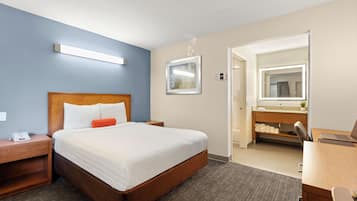 A hotel room with a large bed, a desk, a chair, a nightstand, a bedside table, a mirror, and a bathroom visible through an open door.
