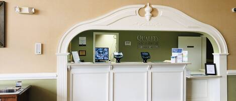 Un comptoir d’accueil surmonté d’une horloge, avec un écran d’ordinateur et une enseigne indiquant « Quality Inn & Suites by Choice Hotels ».