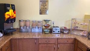 Una estación de desayuno con dispensador de zumos de frutas, varios dispensadores de alimentos, un tostador y una vitrina con bollería.