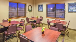 Un salón de conferencias con cuatro mesas y sillas, ventanas amplias con persianas y un arreglo floral en la pared, además de fotografías enmarcadas.