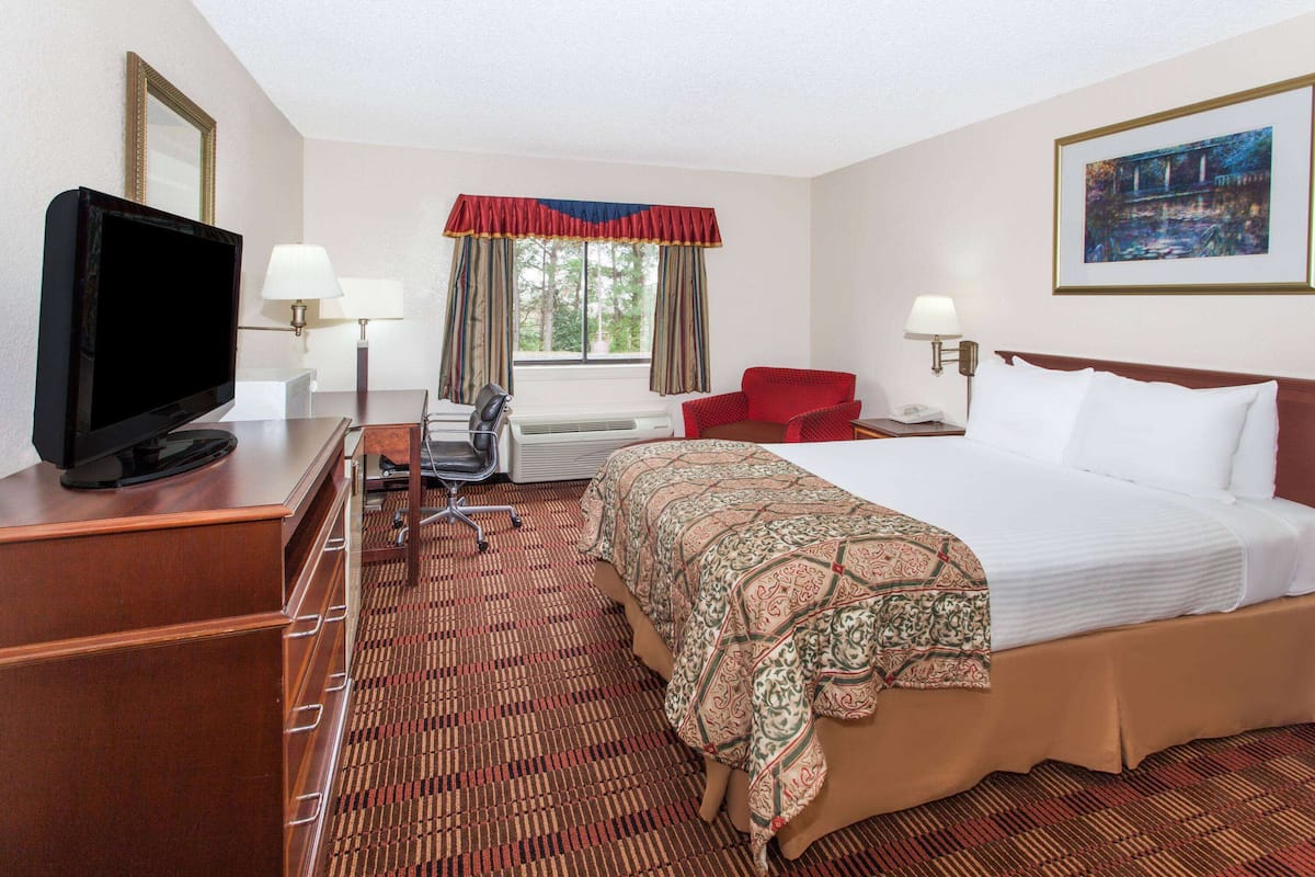 A hotel room with a large bed, a desk with a flat-screen TV, a chair, a desk lamp, and a window with curtains.