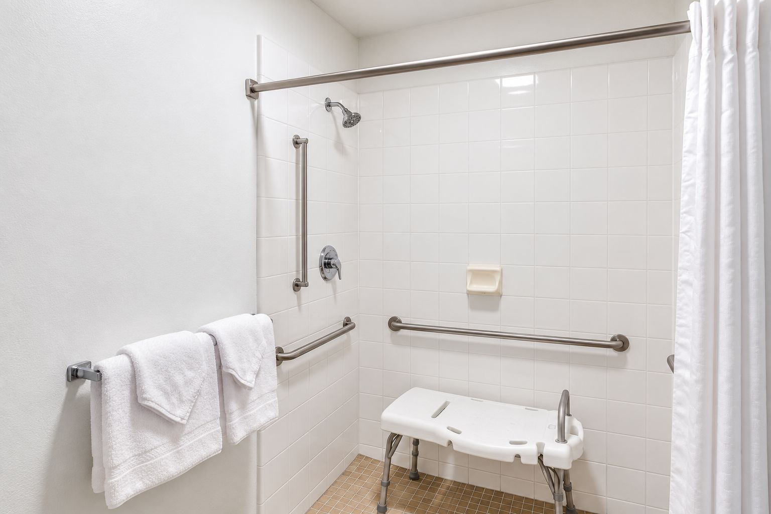 A bathroom with white tiled walls, a shower with a grab bar, a white bench, and a towel rack with towels.