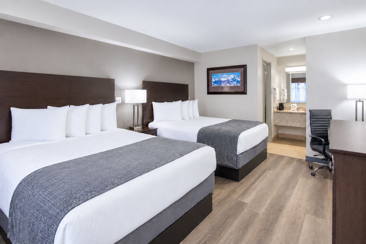 A hotel room with two beds, a desk, a chair, and a bathroom visible through an open door.