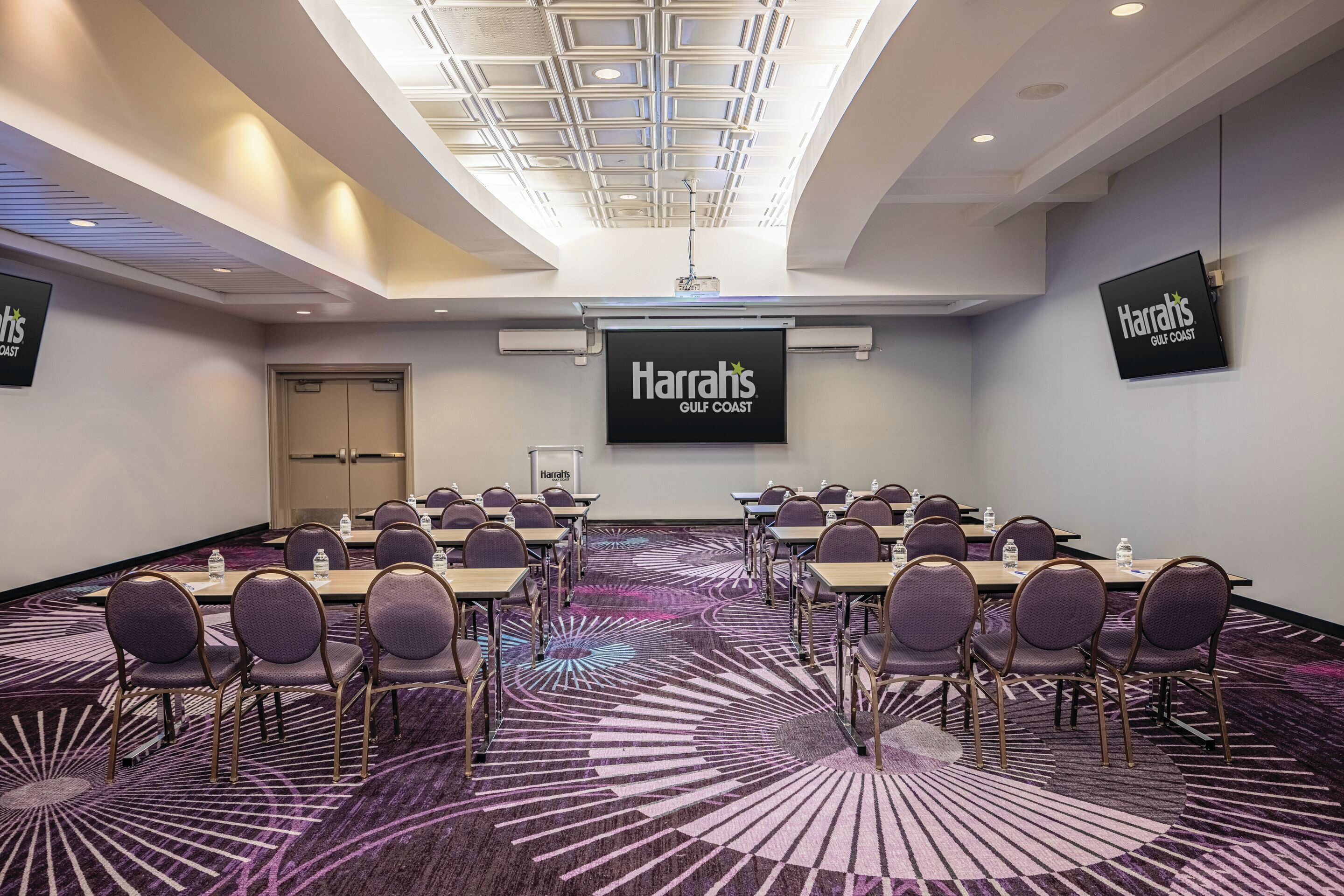 Un salón de conferencias con un gran letrero de Harrah's Gulf Coast, varias mesas con sillas y una alfombra estampada.