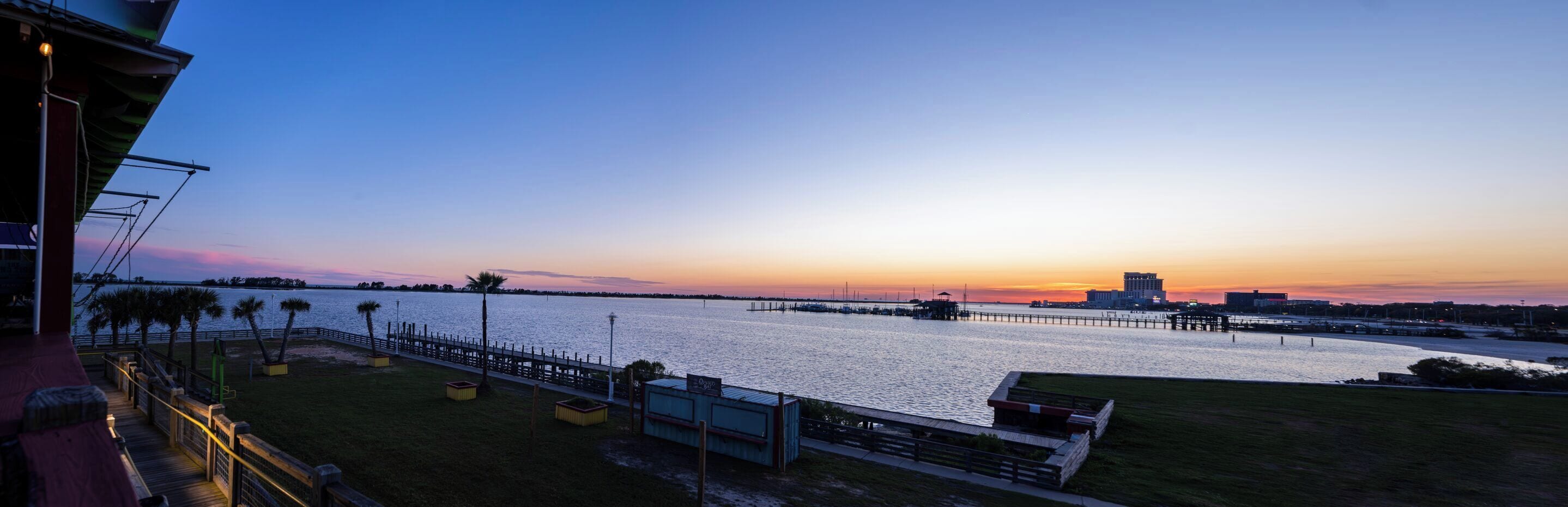 A waterfront view with a wooden deck, palm trees, and a distant city skyline at sunset.