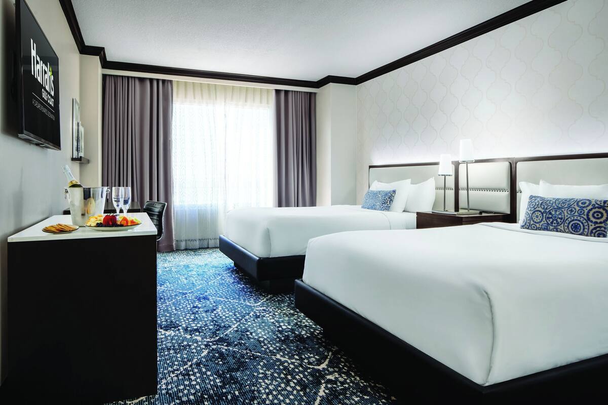 A hotel room with two beds, a desk with breakfast items, a flat-screen TV, and a large window with curtains.