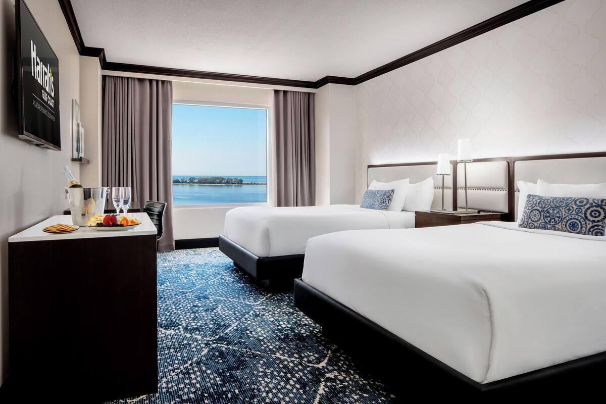 A hotel room with two beds, a desk with a bottle of champagne, and a view of the water.