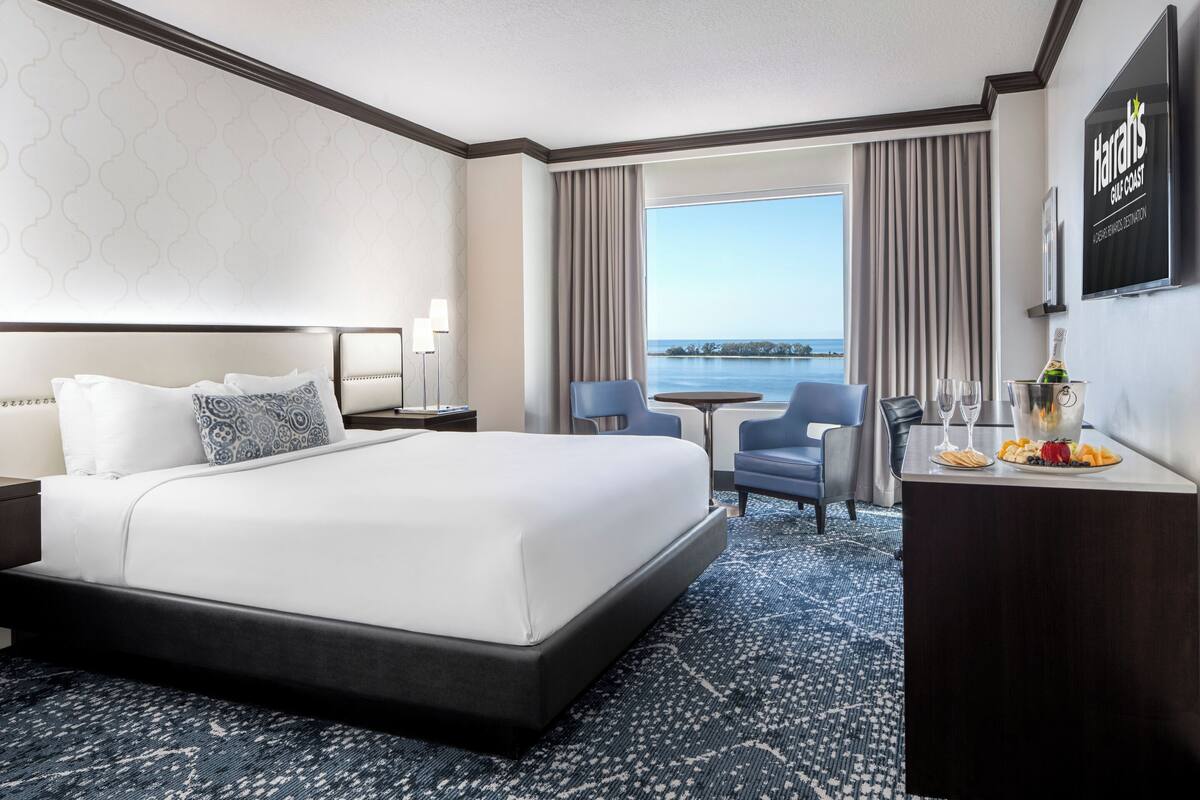 A hotel room with a bed, a desk with a bottle and glasses, a chair, and a view of the sea.