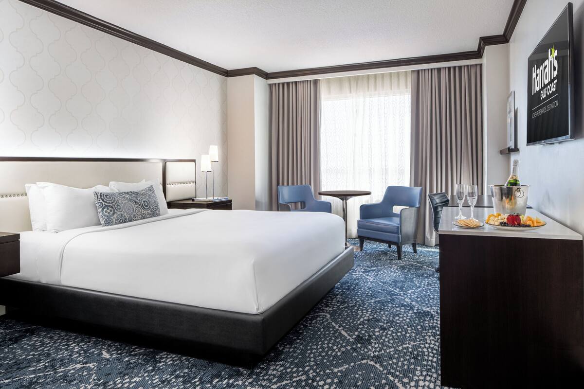 A hotel room with a large bed, a desk with a bottle and glasses, a chair, and a TV on the wall.