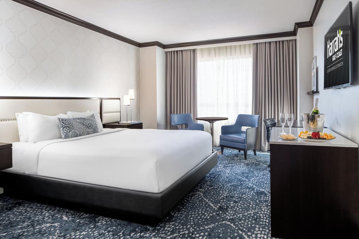A hotel room with a large bed, a desk with a bottle and glasses, a chair, and a TV on the wall.