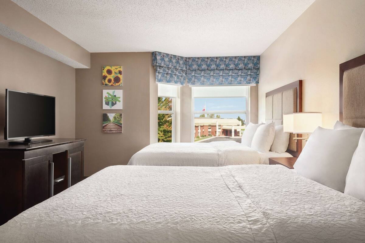 A hotel room with two beds, a TV, and a window with a view of buildings and trees.